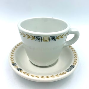 May include: A white ceramic teacup and saucer set with a gold and black geometric design. The design features a repeating pattern of squares and leaves.