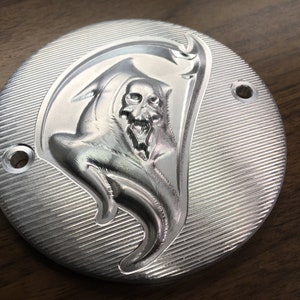 May include: A silver metal round cover with a detailed embossed image of the Grim Reaper holding a scythe. The cover has two small holes on the side.