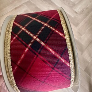 May include: A wide spool of ribbon featuring a red and black plaid pattern with thin gold diagonal lines. The edges of the ribbon are finished with a textured gold metallic trim, providing a distinct border against the fabric spool core.