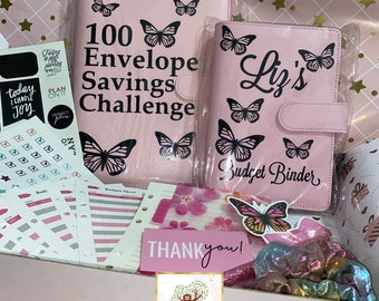 May include: Pink budget binder with butterfly designs and the text "Liz's Budget Binder". A second pink binder with butterfly designs and the text "100 Envelope Savings Challenge" is also visible. The binders are in a white box with other pink and white accessories.