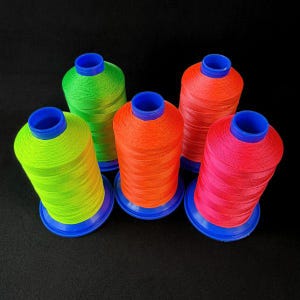Può includere: Cinque rocchetti di filo dai colori vivaci: verde neon, arancione, rosso-arancio e rosa acceso. Ogni rocchetto è cilindrico, con base e parte superiore blu. Il filo è avvolto strettamente, creando un aspetto strutturato su sfondo nero.