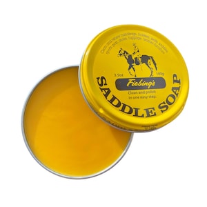 Pode incluir: Uma lata amarela de sabão para sela Fiebing's, um limpador e polidor de couro. A lata tem uma ilustração em preto e branco de uma pessoa a cavalo. O texto na lata diz "Limpe e renove bolsas de mão, coldres, cintos, selas, equipamentos esportivos, sapatos, bagagem, móveis de couro. 3.5oz Fiebing's Saddle Soap 100g Limpe e polir em uma única etapa fácil."