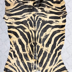 May include: A black and tan zebra hide rug with a natural, distressed look. The hide is large and features a classic zebra stripe pattern.