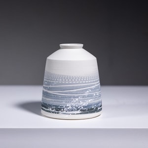 May include: A small white ceramic vase with a blue and white abstract design. The vase has a rounded base and a wide opening.
