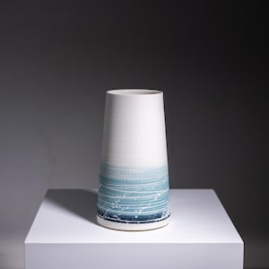 May include: A white ceramic vase with a blue and white abstract design. The vase is tall and narrow, with a rounded base and a flared top.