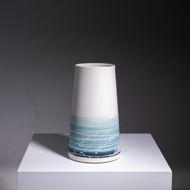 May include: A white ceramic vase with a blue and white abstract design. The vase is tall and narrow, with a rounded base and a flared top.