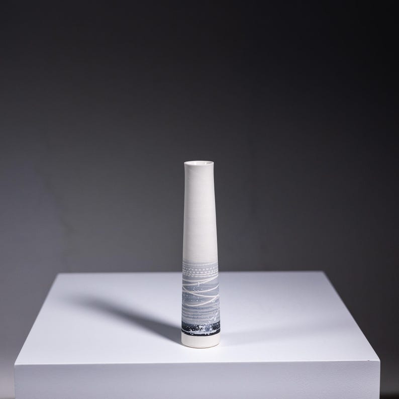 May include: A white ceramic vase with a grey and white geometric pattern. The vase is tall and slender, with a cylindrical shape.