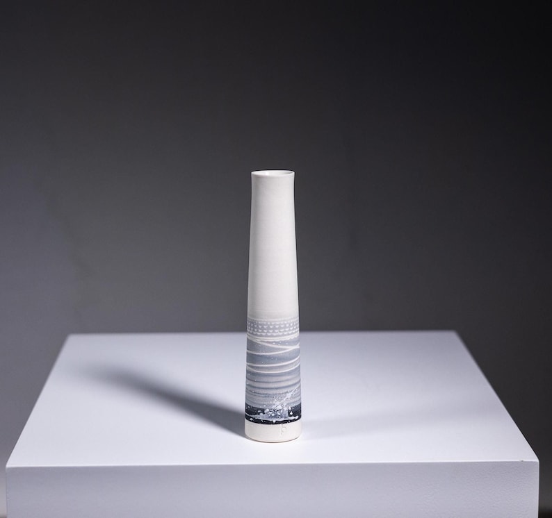 May include: A white ceramic vase with a grey and white abstract design. The vase is tall and slender with a rounded bottom.