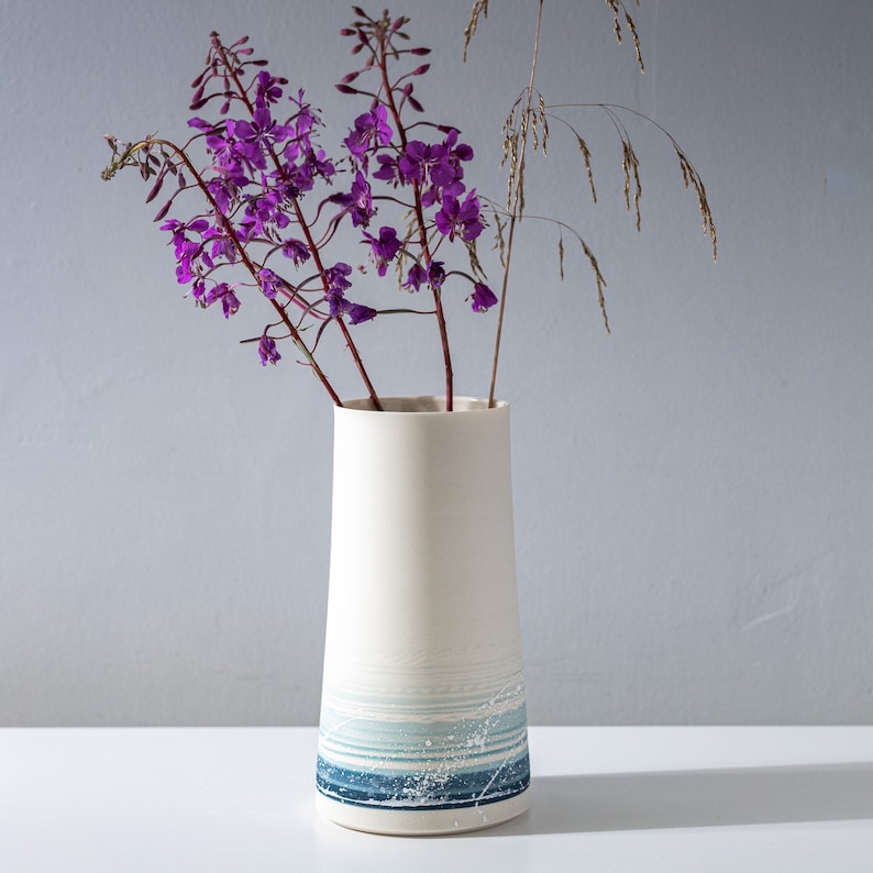May include: A white ceramic vase with a blue and white striped design. The vase is filled with purple flowers.