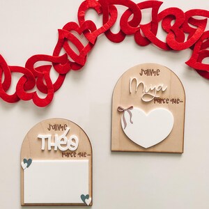 May include: Wooden plaques with arched tops, featuring the phrases "J'aime Thé0 parce que-" and "J'aime Mya parce que-". Each plaque has a white heart or rectangle for personalization. A red felt heart garland is above.