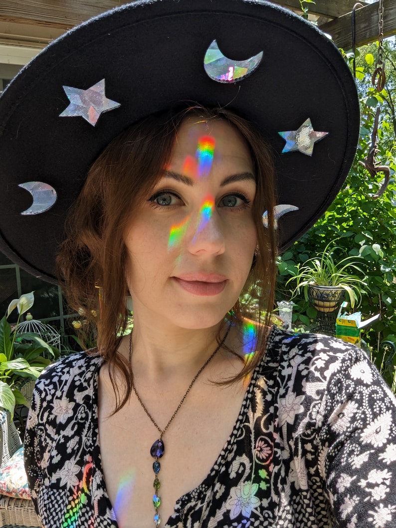May include: A woman wearing a black wide-brimmed hat with iridescent stars and moons. The hat is decorated with holographic accents. She is wearing a black and white floral print top and a long necklace with a large, iridescent gemstone pendant.