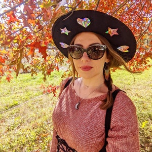 May include: A woman wearing a wide-brimmed black hat with iridescent stars and a heart. She is wearing a red sweater with a black corset and has a silver necklace with a crescent moon pendant. Her hair is pulled back in a braid with beaded earrings.