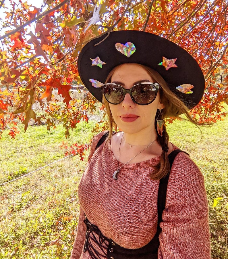 May include: A woman wearing a wide-brimmed black hat with iridescent stars and a heart. She is wearing a red sweater with a black corset and has a silver necklace with a crescent moon pendant. Her hair is pulled back in a braid with beaded earrings.