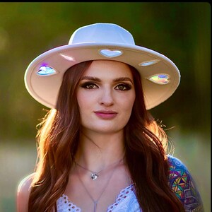 May include: A white wide-brimmed hat with iridescent heart-shaped accents. The hat is being worn by a person with long brown hair.