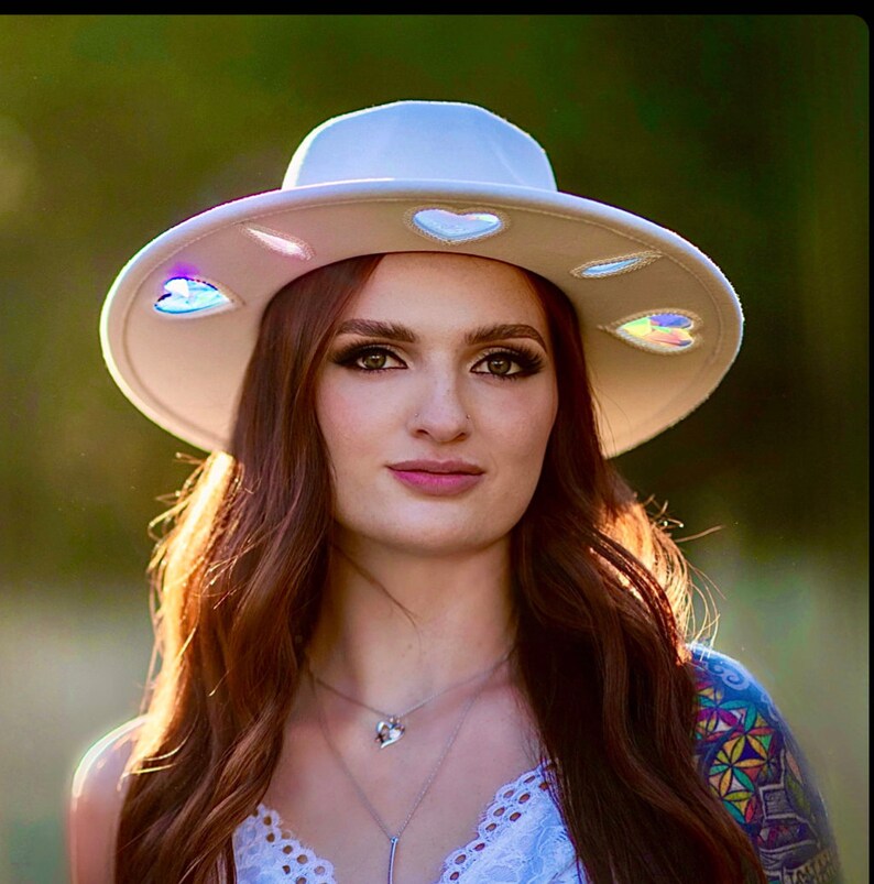 May include: A white wide-brimmed hat with iridescent heart-shaped accents. The hat is being worn by a person with long brown hair.