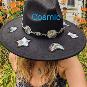 May include: A black felt fedora hat with a wide brim and a silver chain with decorative charms. The hat has a variety of colourful, iridescent patches in the shape of stars and crescent moons. The word "Cosmic" is written in blue on the hat.