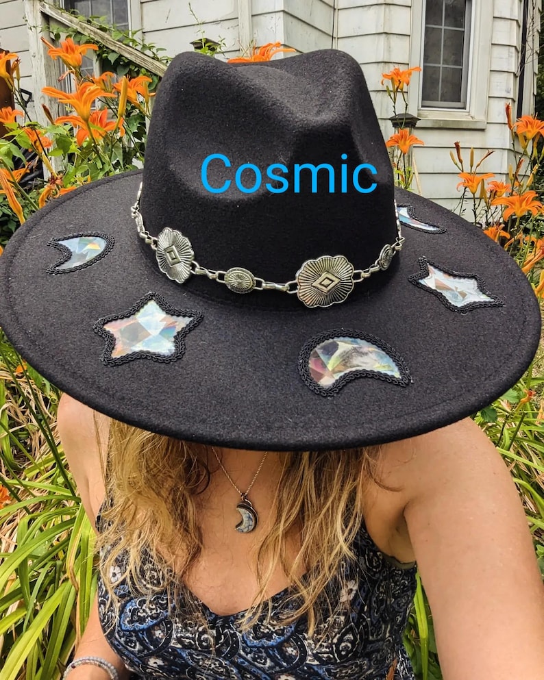May include: A black felt fedora hat with a wide brim and a silver chain with decorative charms. The hat has a variety of colourful, iridescent patches in the shape of stars and crescent moons. The word "Cosmic" is written in blue on the hat.