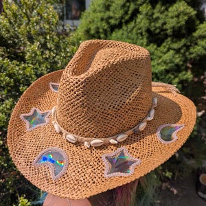 May include: A tan woven straw cowboy hat featuring a band of white cowrie shells around the crown. The wide brim is decorated with iridescent, holographic cutouts in the shapes of stars and a crescent moon, each outlined with light pink stitching.