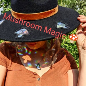May include: A black wide-brimmed hat with a brown velvet band and iridescent mushroom-shaped embellishments. The hat is decorated with the text "Mushroom Magic".