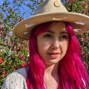 May include: A woman with pink hair is wearing a beige felt hat with a crescent moon and star cutouts. The hat has a gold crescent moon pin on the front.