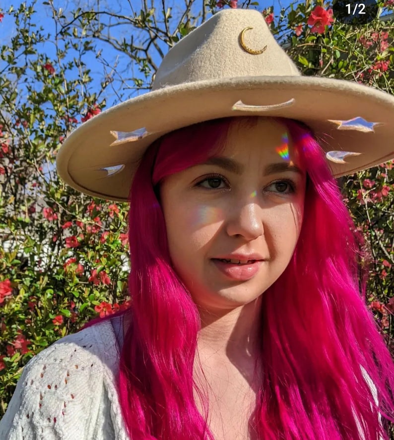 May include: A woman with pink hair is wearing a beige felt hat with a crescent moon and star cutouts. The hat has a gold crescent moon pin on the front.