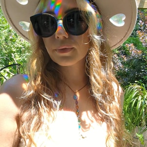 May include: A beige hat with heart-shaped cutouts, worn with black sunglasses. The hat reflects a rainbow of colors. A necklace with colorful beads is also visible. The background includes green foliage and sunlight.