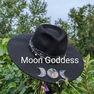 May include: A black wide-brimmed hat with a black ribbon and a triple moon design. The hat is decorated with iridescent sequins and the words "Moon Goddess" are written on the brim.
