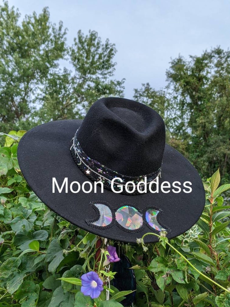 May include: A black wide-brimmed hat with a black ribbon and a triple moon design. The hat is decorated with iridescent sequins and the words "Moon Goddess" are written on the brim.