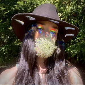 May include: A person wearing a black wide-brimmed hat with a flower covering their mouth. The hat has a decorative design around the brim. The person has long dark hair and is wearing a white shirt.