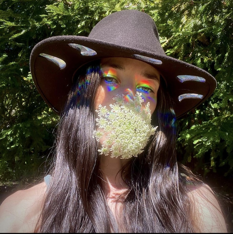 May include: A person wearing a black wide-brimmed hat with a flower covering their mouth. The hat has a decorative design around the brim. The person has long dark hair and is wearing a white shirt.