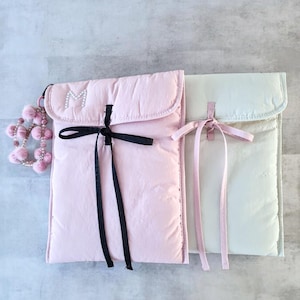 Pode incluir: Duas capas acolchoadas para laptop em cores pastel. Uma é rosa claro com fecho de fita preta e uma letra "M" com joias. A outra é verde pálido com fecho de fita rosa. Um pingente de pompom rosa pende da capa rosa.