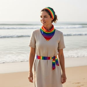 Puede incluir: Una mujer con una diadema, bufanda y cinturón a rayas arcoíris. Los accesorios presentan un vibrante espectro de colores. Lleva un vestido beige. La escena está ambientada en una playa con el océano de fondo.