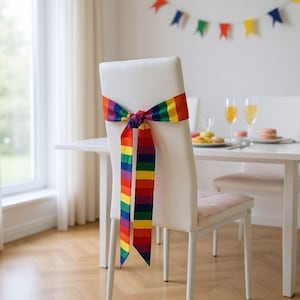 Könnte beinhalten: Ein weißer Stuhl mit einem Regenbogenband, das um die Rückenlehne gebunden ist. Das Band zeigt Streifen in Rot, Orange, Gelb, Grün, Blau und Lila. Der Stuhl ist Teil einer Essgruppe, mit einem Tisch und anderen Stühlen im Hintergrund.