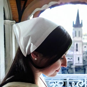 May include: A white head covering, possibly a bonnet or scarf, is worn on the head. The head covering is made of a crisp, white fabric. The person wearing the head covering has dark hair and a pearl earring. The background includes a building with a tall spire.