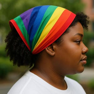 Può includere: Una fascia a righe arcobaleno con bande rosse, arancioni, gialle, verdi, blu e viola. La fascia è indossata sulla testa. Lo sfondo è sfocato, con fogliame verde visibile.