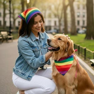 Puede incluir: Una mujer y un perro golden retriever en un parque. La mujer lleva una chaqueta vaquera, pantalones blancos y una diadema arcoíris. El perro tiene un pañuelo arcoíris. La mujer acaricia al perro.