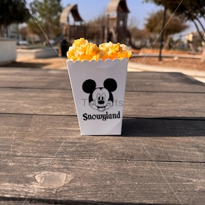 May include: A white popcorn container featuring a Mickey Mouse design and the text "Snowland" on the front. The container is filled with yellow popcorn.