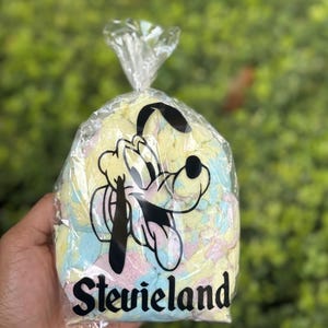 Peut inclure: Un sac en plastique transparent rempli de guimauves colorées. Le sac présente un dessin de personnage de dessin animé en noir et blanc et le mot "Stevieland" en gras, en noir. Les guimauves sont jaune pastel, bleu et rose.