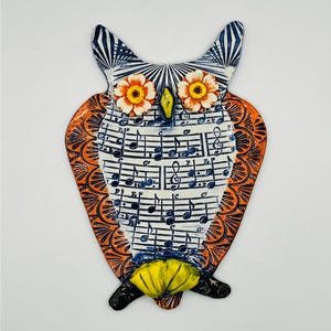 May include: A ceramic owl wall plaque featuring a white chest decorated with black musical notes and staves. The owl has orange patterned wings, two raised orange and white flower-shaped eyes, a yellow beak, and yellow feet at the base.