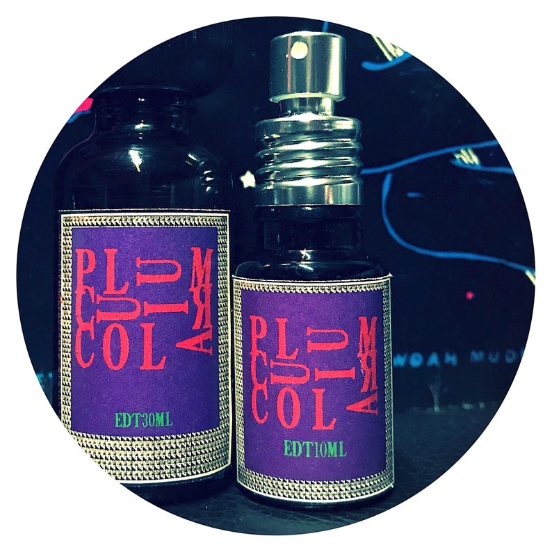 May include: Two glass bottles with purple labels and silver caps. The labels have the text "PLUM COLA" in red letters. The smaller bottle says "EDT 10ML" and the larger bottle says "EDT 30ML".