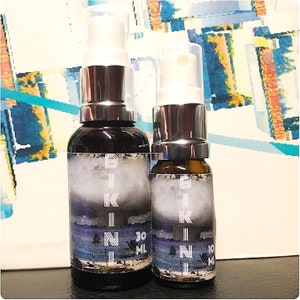 Könnte beinhalten: Zwei Glasflaschen mit silbernen Sprühköpfen und Etiketten mit der Aufschrift "BIKINI" und einer Strandszene. Die größere Flasche ist 30 ml und die kleinere Flasche ist 10 ml.