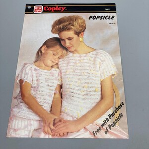 May include: A vintage Copley pattern titled "Popsicle" featuring a mother and child wearing matching crocheted tops. The tops are white with pink and yellow accents. The pattern is 26.44 inches in length.