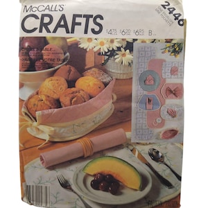 May include: A McCall's Crafts sewing pattern envelope 2446 featuring instructions for fabric table accessories. The cover displays a quilted bread basket, a rolled napkin with a ring, and various placemats with floral patterns and pink accents.