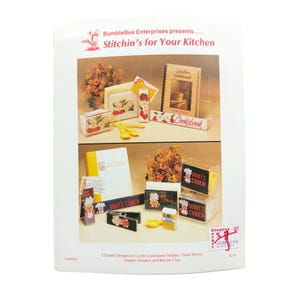 May include: A craft pattern leaflet titled Stitchin's for Your Kitchen by BumbleBee Enterprises. It displays charted designs for lucite cookbook holders, index boxes, napkin holders, and recipe clips featuring floral and chef motifs.