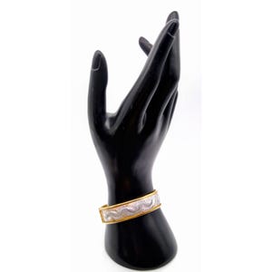 May include: A gold-coloured metal bangle bracelet with a silver-coloured, patterned centre band. The bracelet is displayed on a black hand-shaped jewellery holder against a white background. The bracelet has a hinged clasp.