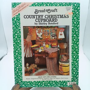 Pode incluir: Uma capa de livro para um projeto de artesanato intitulado "Country Christmas Cupboard" de Shirley Botsford. A capa apresenta uma ilustração colorida de uma cozinha de estilo rústico com um armário de Natal decorado, figuras de pão de gengibre e outras decorações festivas.