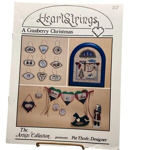 May include: A page from "HeartStrings" featuring a Christmas-themed cross-stitch pattern collection. The designs include ornaments, a framed scene, and a banner. The text reads "A Cranberry Christmas" and "The Artists Collection presents Pat Thode, Designer."
