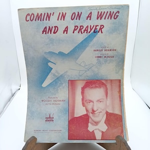 May include: A vintage sheet music cover for the song "Comin' In On A Wing And A Prayer" featuring a blue and white illustration of an aeroplane and a photo of Woody Herman. The cover is printed on light blue paper with red and black text.
