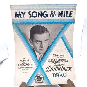 Puede incluir: Una portada de partitura vintage para la canción "My Song of the Nile" de la película First National Vitaphone Picture "Drag" protagonizada por Richard Barthelmess. La portada presenta una foto en blanco y negro de Richard Barthelmess y el texto "My Song of the Nile" en letras azules y blancas.