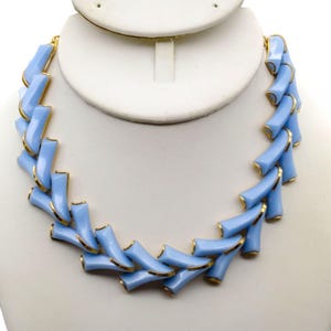May include: A necklace featuring a series of light blue, curved, cylindrical segments linked together. Each segment is accented with a thin gold-toned metal trim along the edges, creating a repeating pattern that forms a semi-circular shape.
